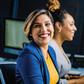 Smiling woman with short hair in a blue blazer and yellow shirt. Smiling woman slightly out of focus next to her.